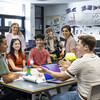 Wide shot of a group of students and their teacher gathered around a table in a technology classroom.